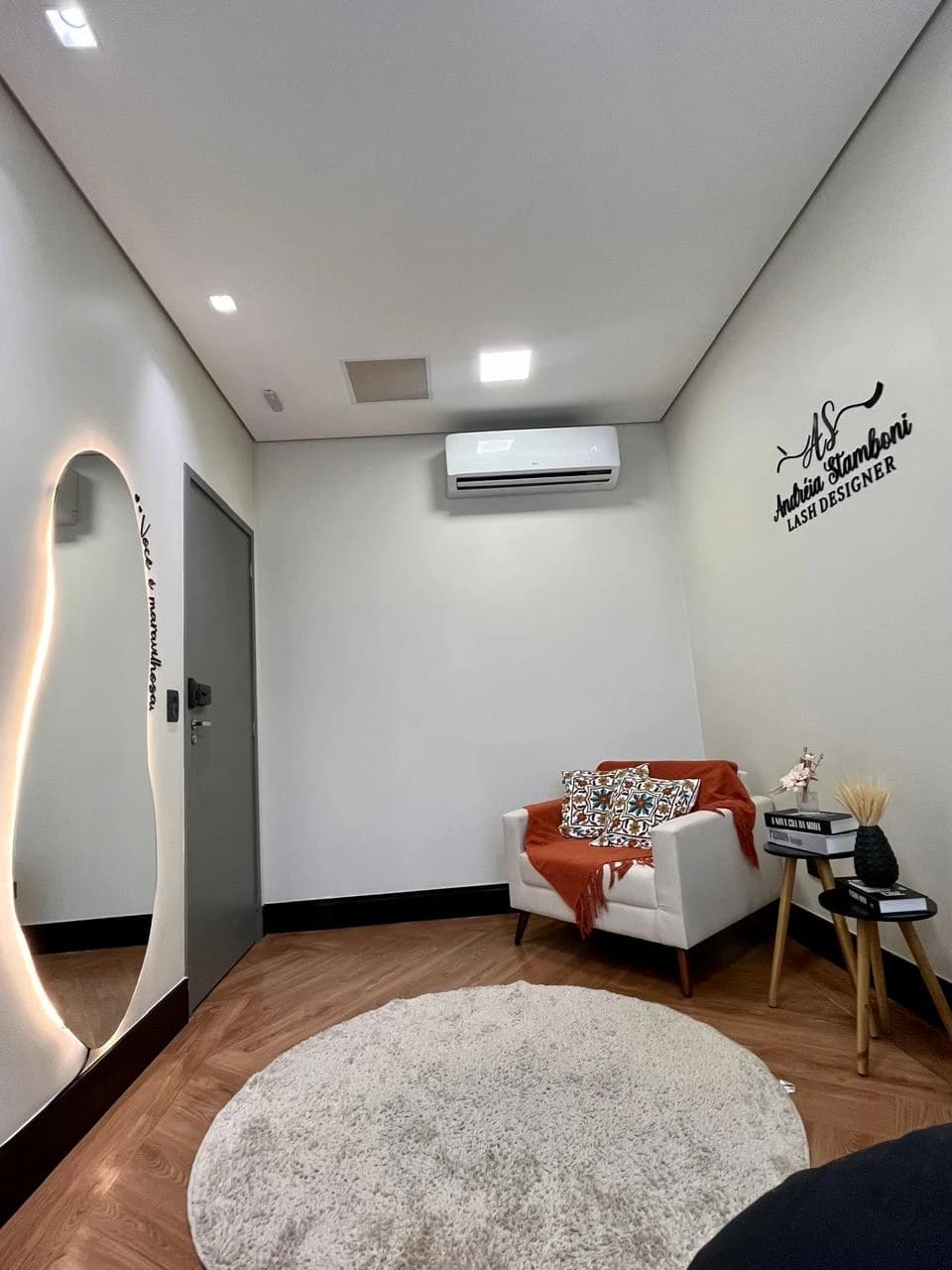 Consultório com o logo Andréia Stamboni Lash Designer na parede, poltrona branca com manta laranja e tapete redondo